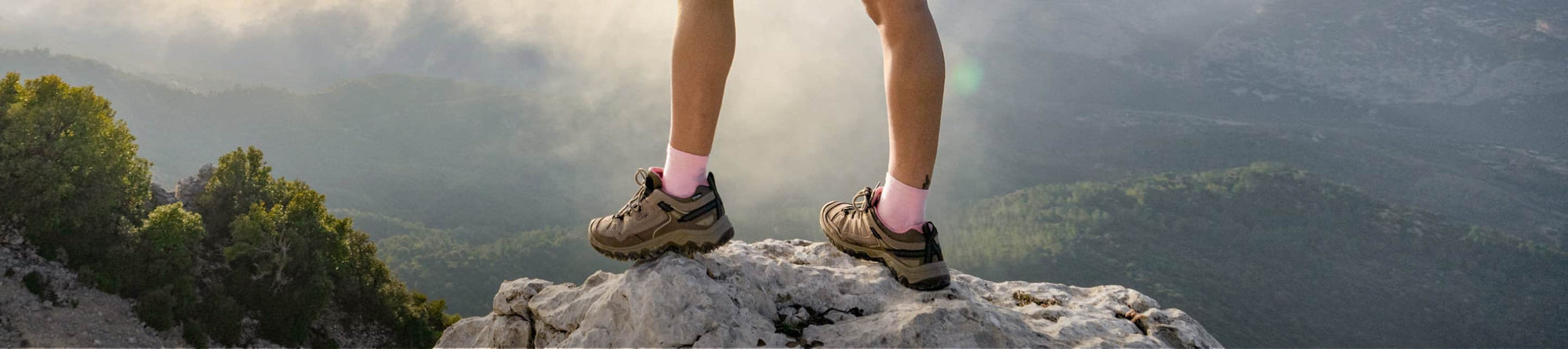 Frau in kurzer Hose steht in hellbraunen, knöchelhohen Wanderschuhen auf einem Felsen mit Aussicht auf eine bergige Waldlandschaft.