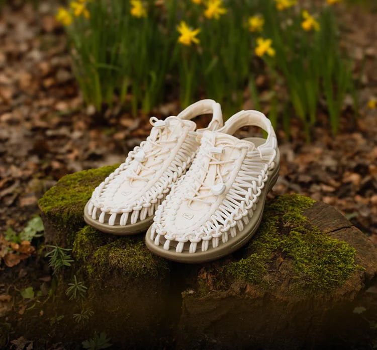 Ein Paar cremefarbene UNEEK-Schnürschuhe mit brauner Sohle ist auf einem mit Moos bewachsenem Stein platziert, der sich in einem Waldgebiet mit Blumen befindet. 